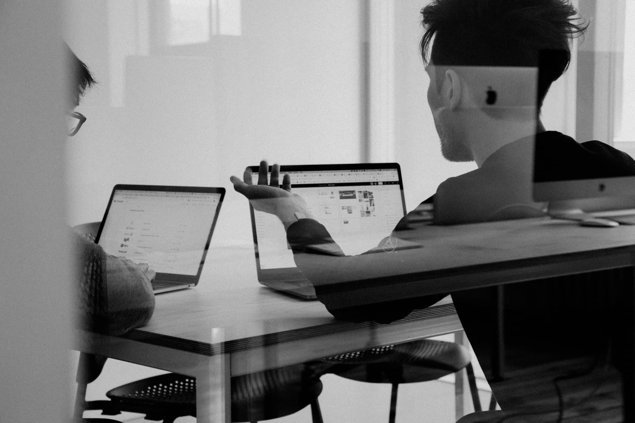 Duas pessoas conversando em uma mesa de trabalho com notebooks abertos, vistas através do vidro da sala